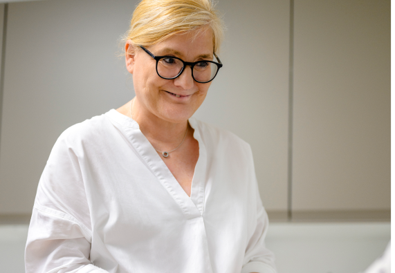 Blonde Person mit Brille lächelt, hält Smartphone wohl zum Herz-Check-Up planen, trägt weiße Bluse, steht vor grauen Schränken.