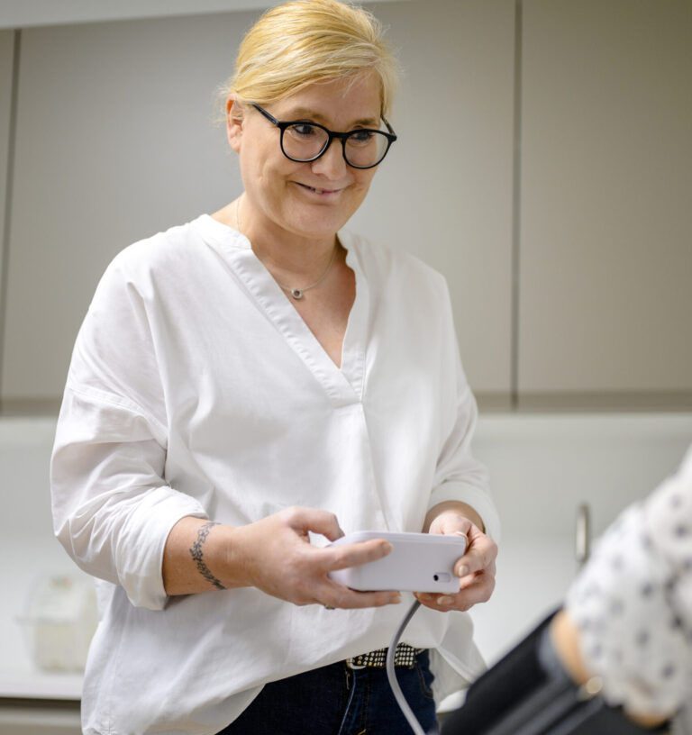 Person mit Brille und weißer Bluse lächelt mit Smartphone im Raum, während jemand auf Herz-Check-Up vorbereitet wird.