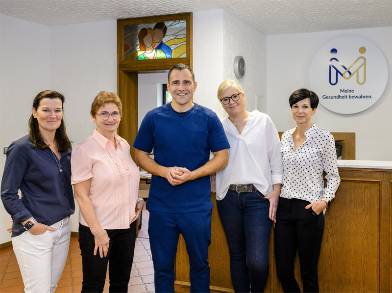 Fünf Erwachsene, darunter einer in blauem Kittel, lächeln in einer Arztpraxis mit einem Schild über Fußschmerztherapie.