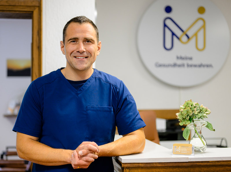 Lächelnder Mann in blauem Kittel an der Rezeption; Visitenkartenhalter, Pflanze und Schild "Stoßwellentherapie Fuß" im Hintergrund.