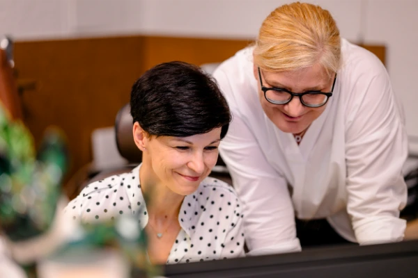 Zwei Frauen im Büro schauen gemeinsam am Computer Informationen zu Hyaluronspritze Knie nach.