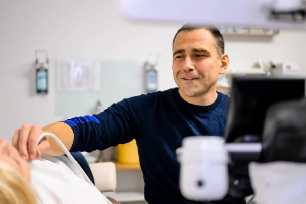 Ein lächelnder Mann sitzt neben einer Person, die sich einer Ultraschalluntersuchung der Schulter unterzieht, mit medizinischen Geräten und Monitoren im Hintergrund.