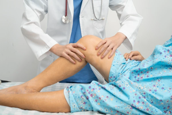 Ein Arzt im weißen Kittel untersucht das Knie eines Patienten, der in einem blauen geblümten Kittel auf dem Untersuchungstisch liegt.